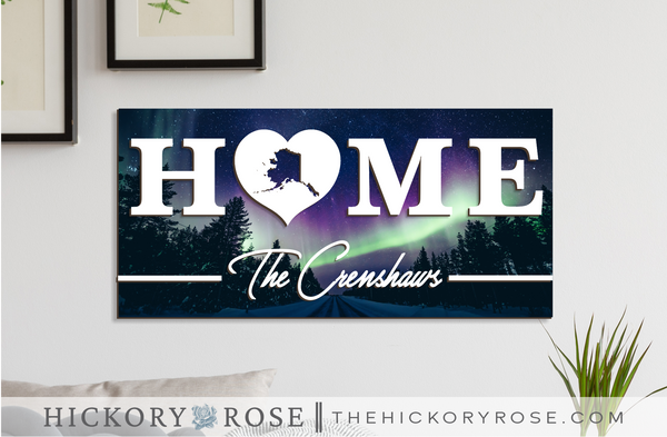 Alaska "HOME" sign with a background of the northern lights over a dark snowy road.
